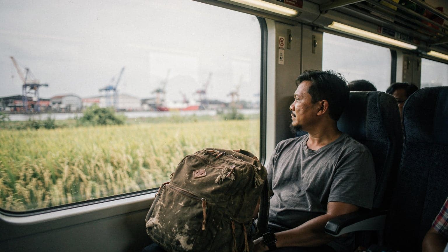 En reisende sitter ved vinduet på et tog fra Yogyakarta mot Surabaya, ser ut over rismarker og en havn i det fjerne, bag på setet ved siden av.