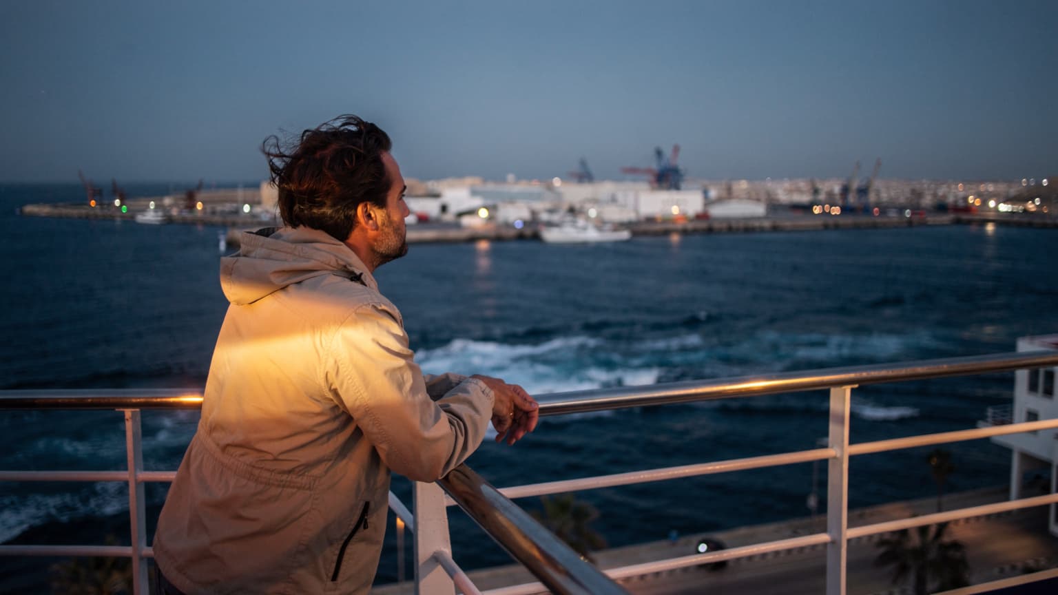 Une personne, le vent dans les cheveux, accoudée à la rambarde d'un ferry quittant le port de La Goulette, Tunis, au crépuscule. Les lumières de la ville brillent à l'arrière-plan.