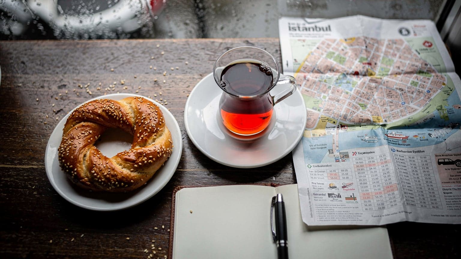 Tavolo di un caffè a Kadıköy con tè turco, simit e una mappa di Istanbul, luce grigia di pioggia dalla finestra