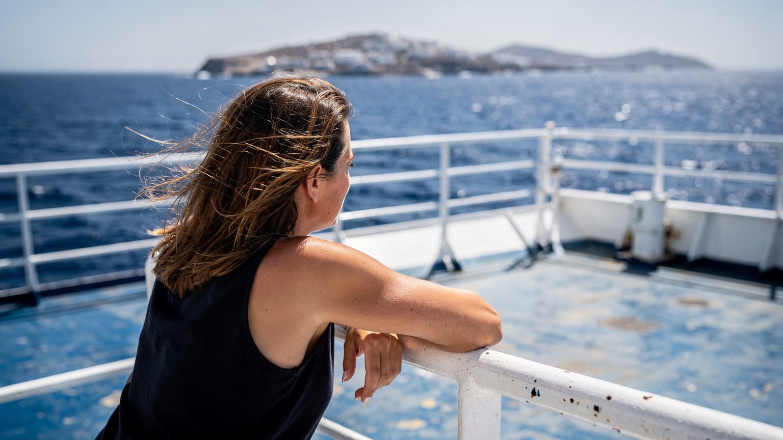 Γυναίκα ακουμπισμένη στην κουπαστή του πλοίου για τη Μύκονο, με τον άνεμο στα μαλλιά και τις Κυκλάδες στο βάθος, κάτω από τον μεσημεριανό ήλιο.