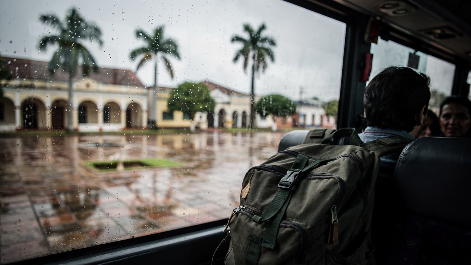 Ventana de bus en ruta hacia Asunción con lluvia ligera, una plaza andaluza de fondo y una mochila en el asiento contiguo