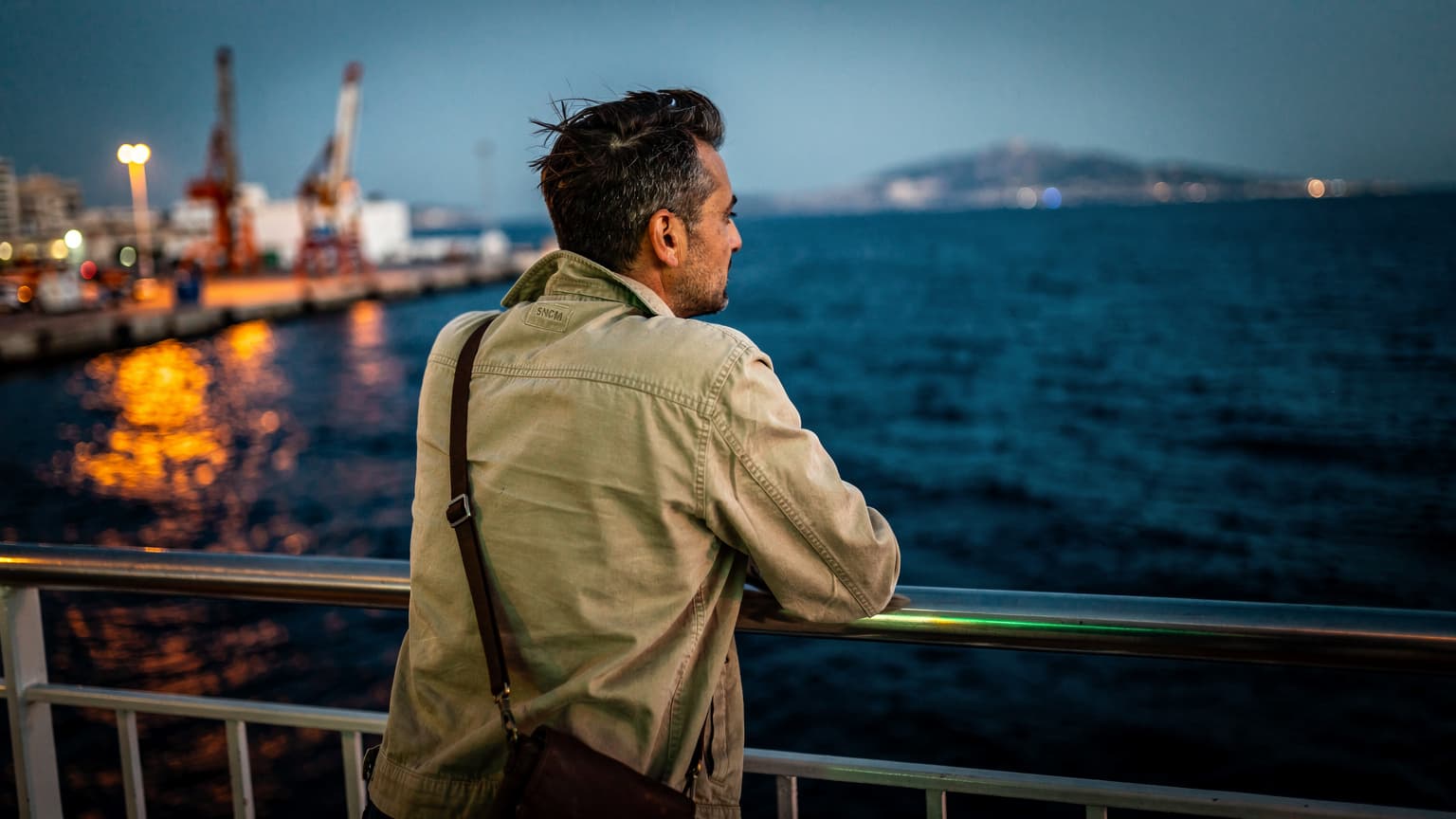 Voyageur appuyé sur le bastingage d'un ferry au départ de Marseille, côte tunisienne au loin, lumière du soir mêlant tungstène et néon