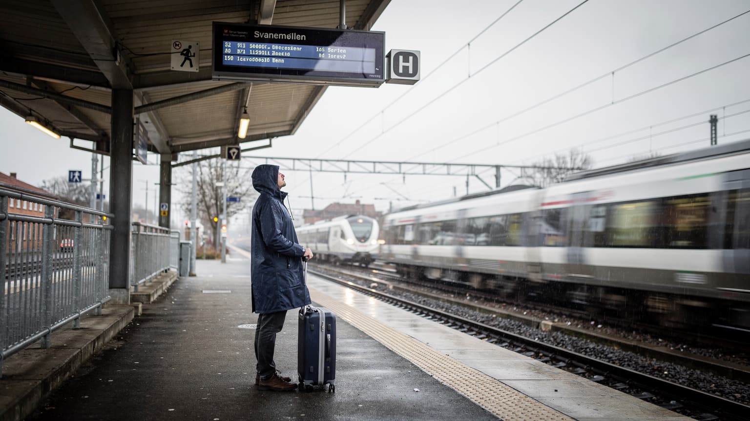 En rejsende på en forstadsbanegård i København læser afgangstavlen i gråt regnvejr, med kuffert ved fødderne