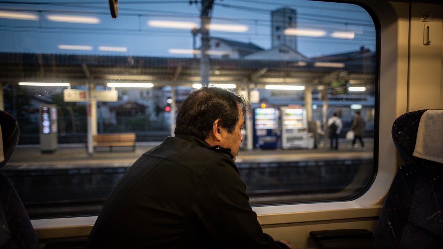 ภาพคนเดินทางมองออกไปนอกหน้าต่างรถไฟในช่วงเวลาสนธยาที่สถานีรถไฟชานเมืองในญี่ปุ่น มีแสงไฟจากชานชาลาสะท้อนบนกระจก