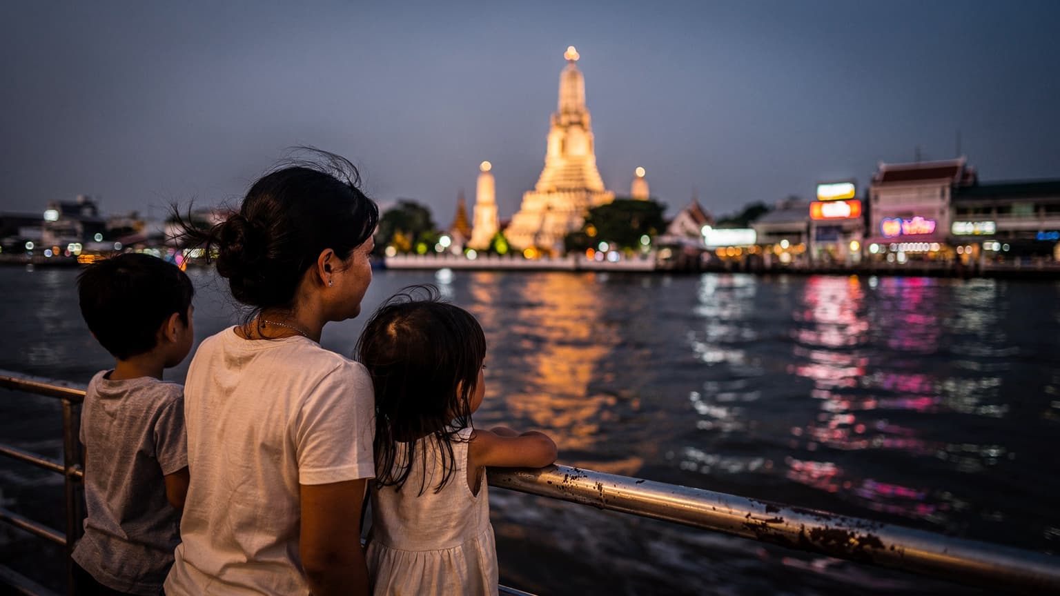 ครอบครัวนั่งเรือข้ามฟากในยามเย็น มองเห็นวัดอรุณและแสงไฟนีออนสะท้อนน้ำ ขณะกำลังมุ่งหน้าไปเที่ยวเทศกาลออกพรรษา