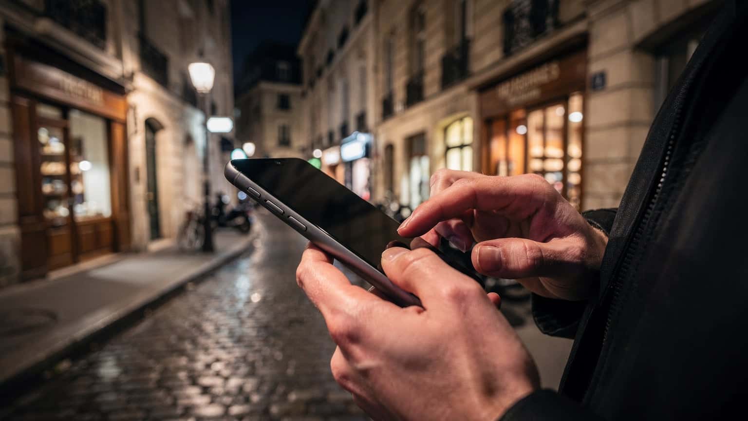 Късно вечер, в тясна павирана уличка в Париж, човек държи смартфон в ръцете си, докато екранът е обърнат далеч от камерата. Неоновите светлини на близките магазини и топлите жълти светлини от прозорците хвърлят сложни сенки върху старите каменни стени, създавайки интимна и загадъчна атмосфера.