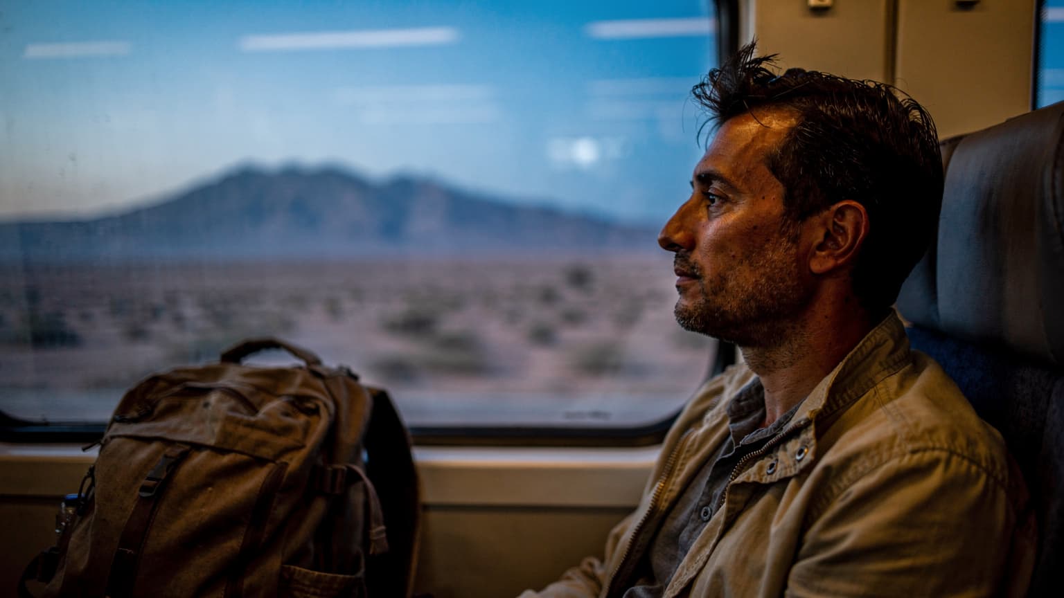 Un voyageur regarde le paysage marocain défiler par la fenêtre d'un train, sac posé sur le siège voisin, à l'heure bleue