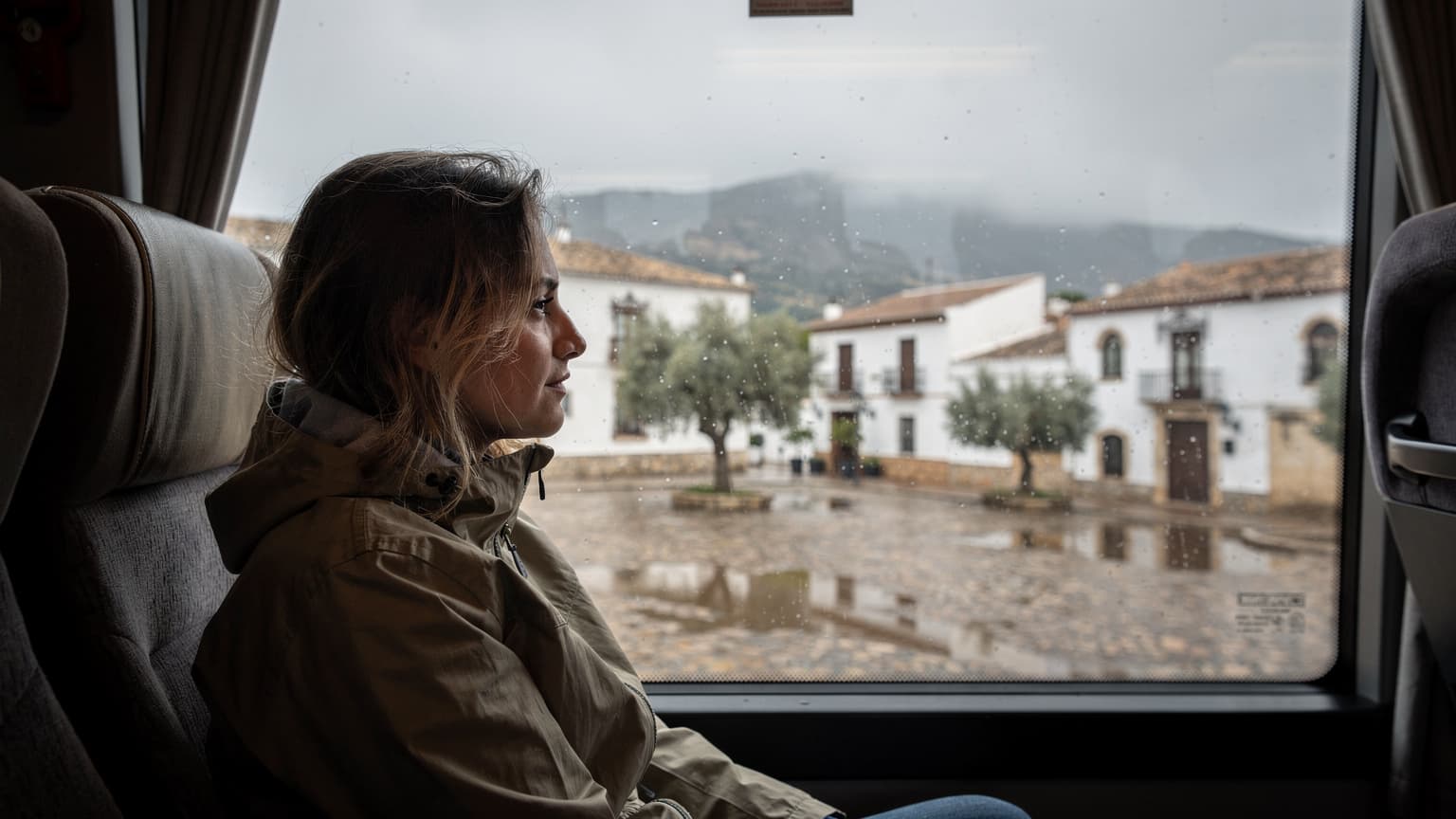 Una viajera mira por la ventana de un autobús bajo la lluvia, observando una plaza de pueblo andaluz con fachadas blancas y tejados ocres que se reflejan en el cristal húmedo.