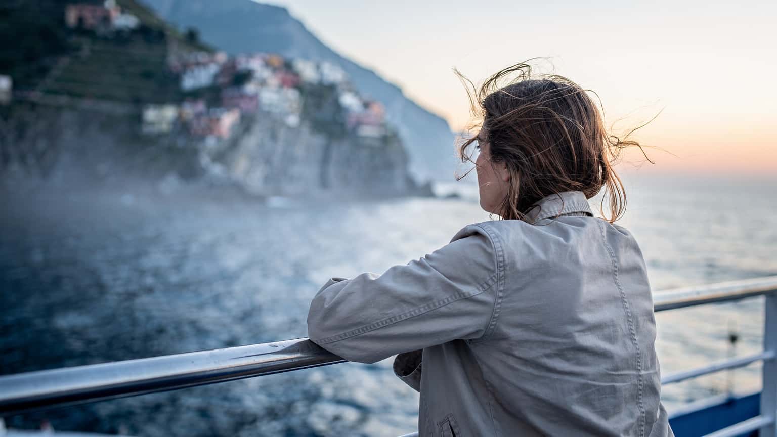 Viajante encostado à amurada de um ferry, com o vento no cabelo, ao amanhecer, com a costa italiana ao fundo, vista das traseiras, luz azul fria.