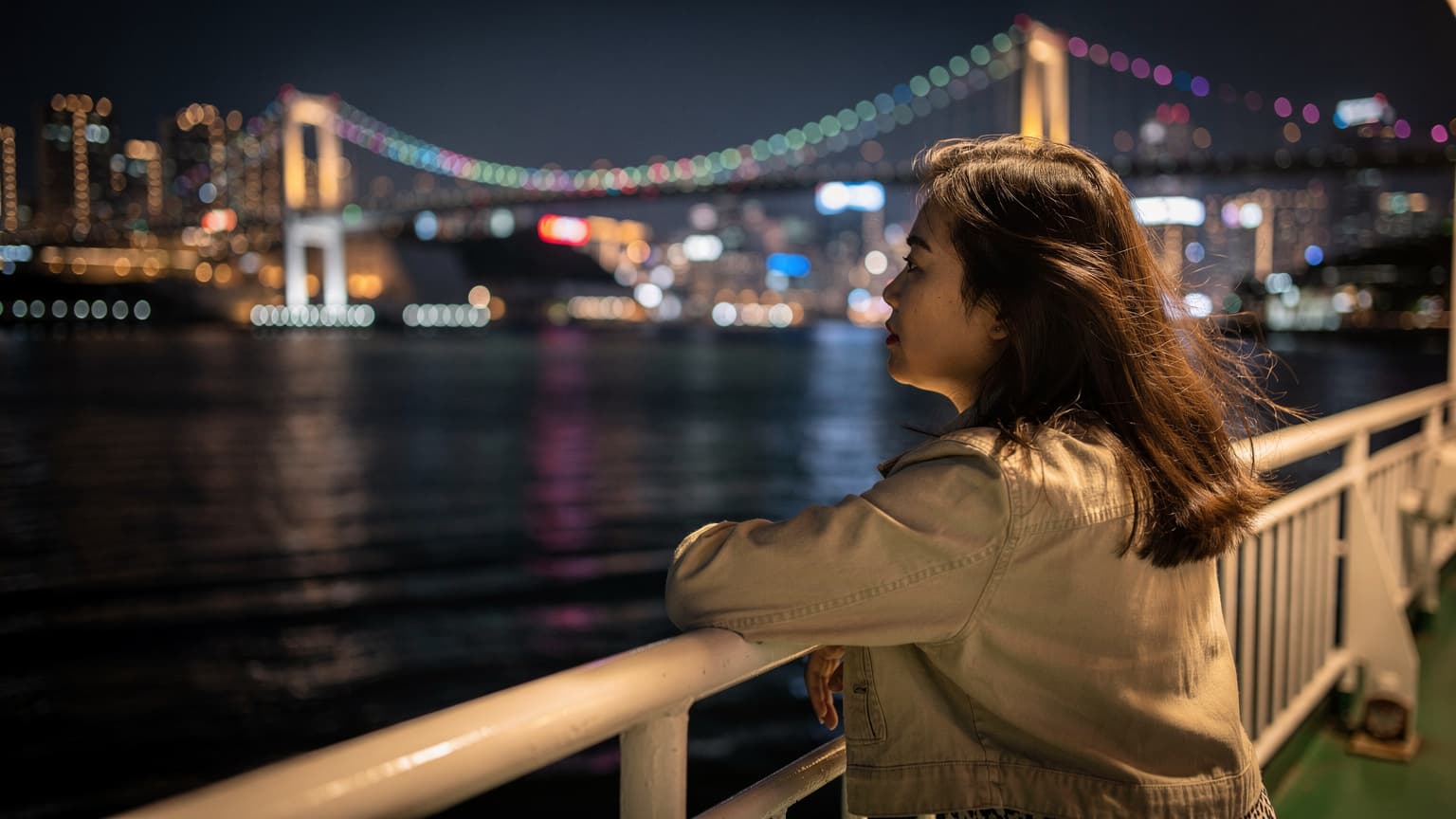 Một người phụ nữ Việt Nam đứng trên boong tàu phà ở Busan vào ban đêm, tay vịn lan can, mái tóc bay trong gió, phía sau là ánh đèn rực rỡ của cầu Gwangan.