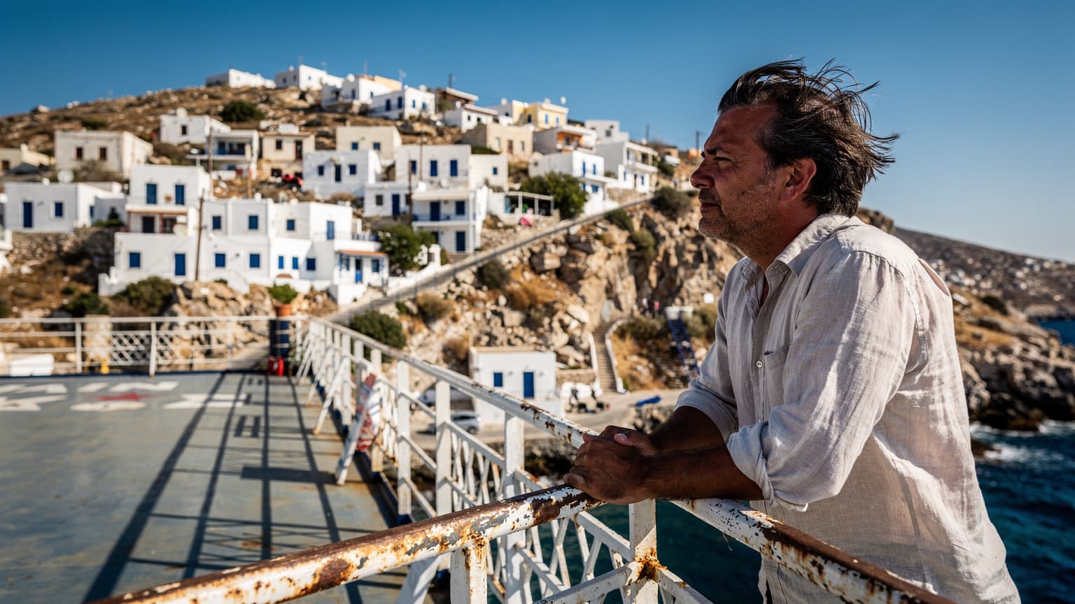 Επιβάτης ακουμπισμένος στο κάγκελο του πλοίου Blue Star στο Αιγαίο, με τον άνεμο στα μαλλιά και την ακτογραμμή της Τήνου στο βάθος, μεσημεριανό φως.