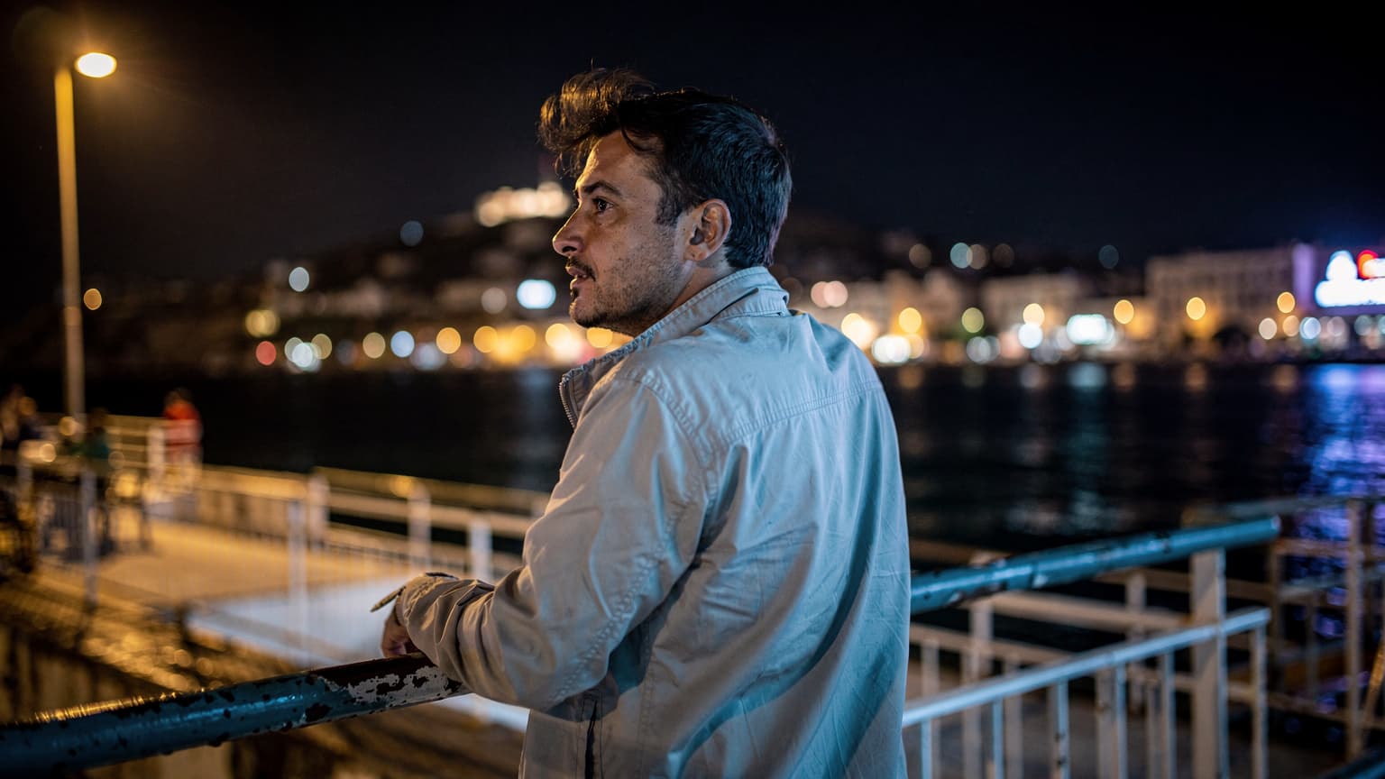 Voyageur adossé au bastingage d'un ferry au port de La Goulette, nuit, lumière tungstène et néon, côte tunisienne en arrière-plan