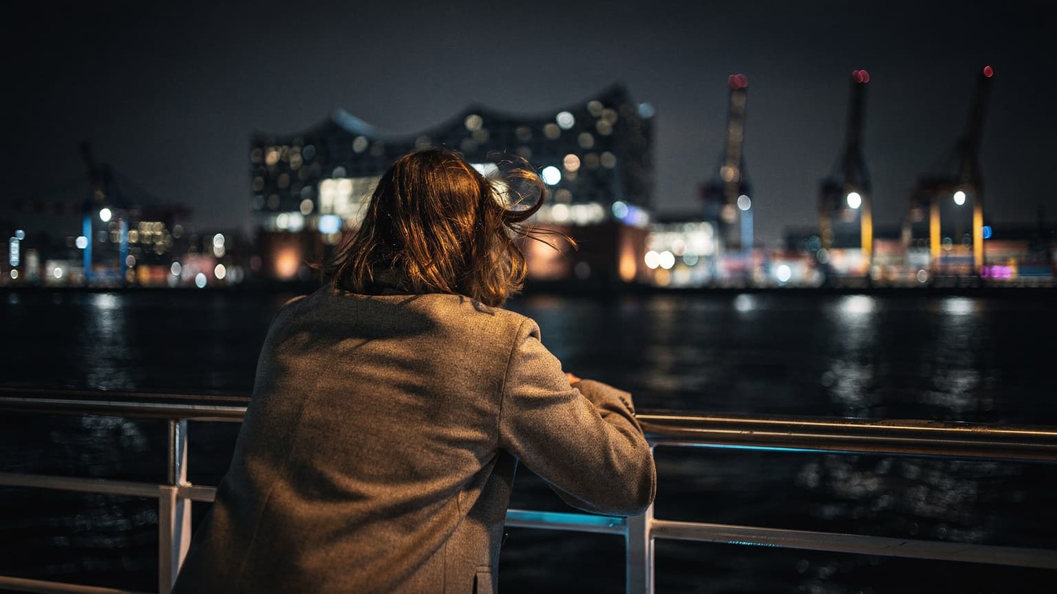 Eine Person lehnt nachts auf dem Deck einer Fähre im Hamburger Hafen, der Wind zerzaust ihr Haar, während im Hintergrund die beleuchtete Elbphilharmonie und Hafenkräne vorbeiziehen.