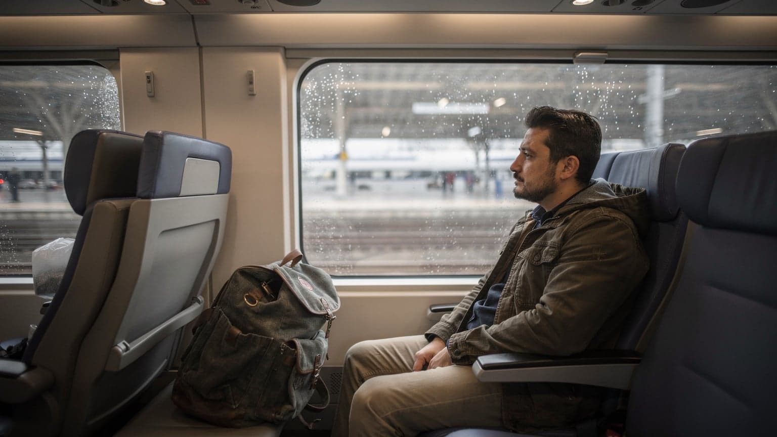 Ankara YHT peronunda bir tren penceresinden dışarıya, yağmurlu gri bir havada bulanıklaşan manzarayı izleyen bir gezgin. Koltuğunda sırt çantası duruyor.