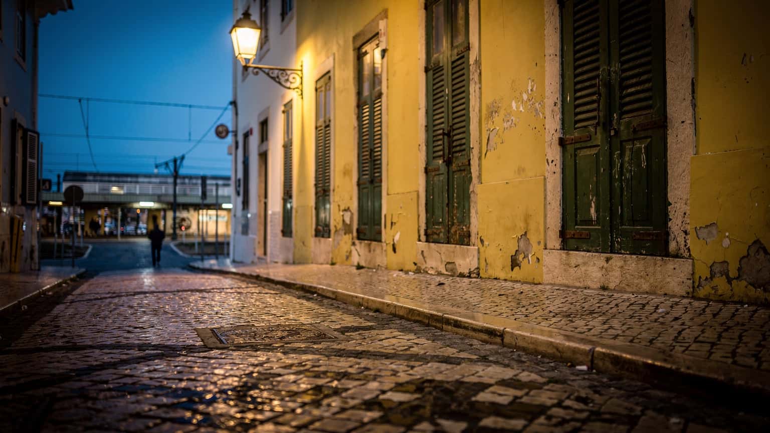 Rua de calçada portuguesa ao entardecer, junto à estação de comboios de Lisboa, com luz azul e persianas verdes fechadas.
