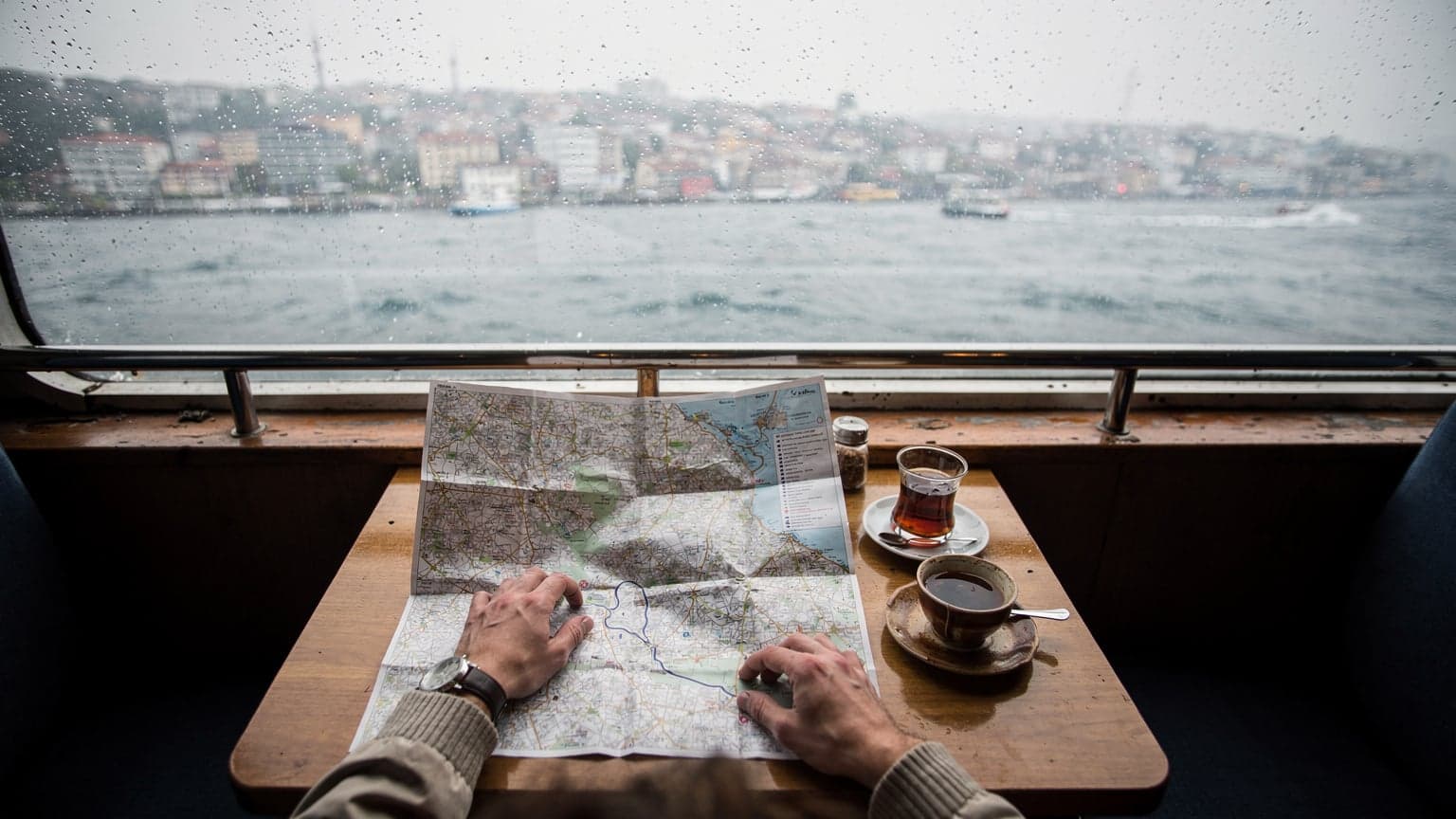 Un caffè mattutino su un traghetto che attraversa il Bosforo a Istanbul, con un bicchiere di tè turco, un simit e una mappa cartacea del quartiere di Karaköy appoggiati su un tavolo di legno bagnato dalla pioggia leggera.