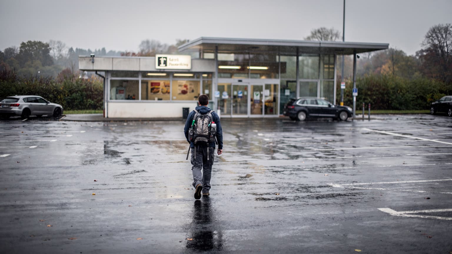 Ein nasser, grauer Autobahnraststättenparkplatz in Deutschland bei Regen, mit einem Schild, das auf einen Imbiss hinweist.