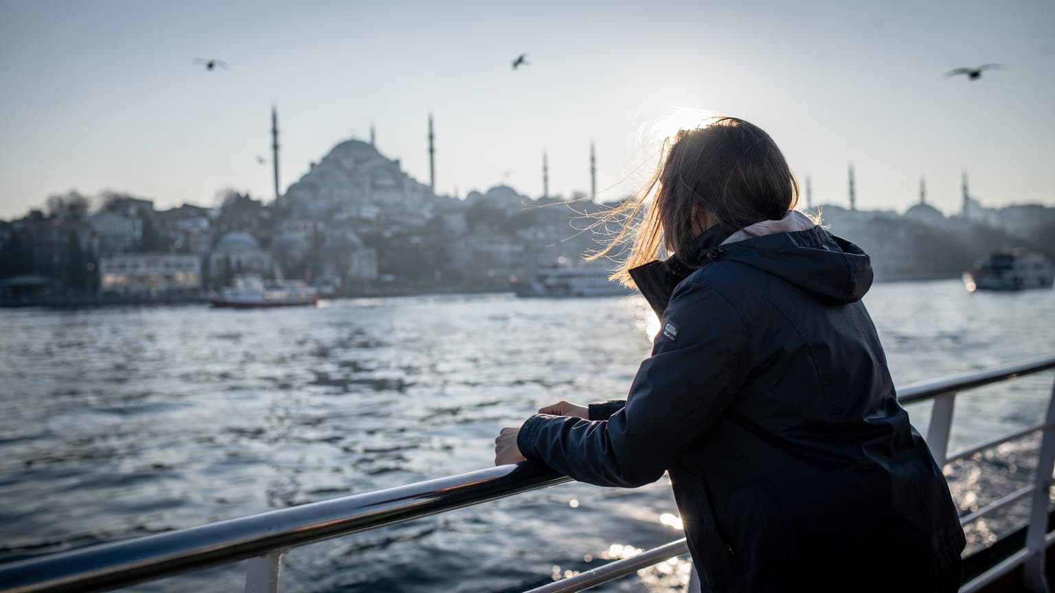 Rejsende læner sig over gelænderet på en færge ved havn i Istanbul, morgenlys over dækket, ingen telefon i hånden