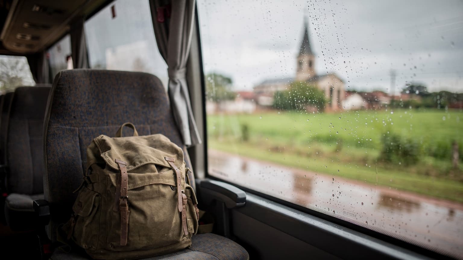 Ventana de bus rural en Paraguay con lluvia, paisaje verde y una mochila en el asiento, luz gris de tarde