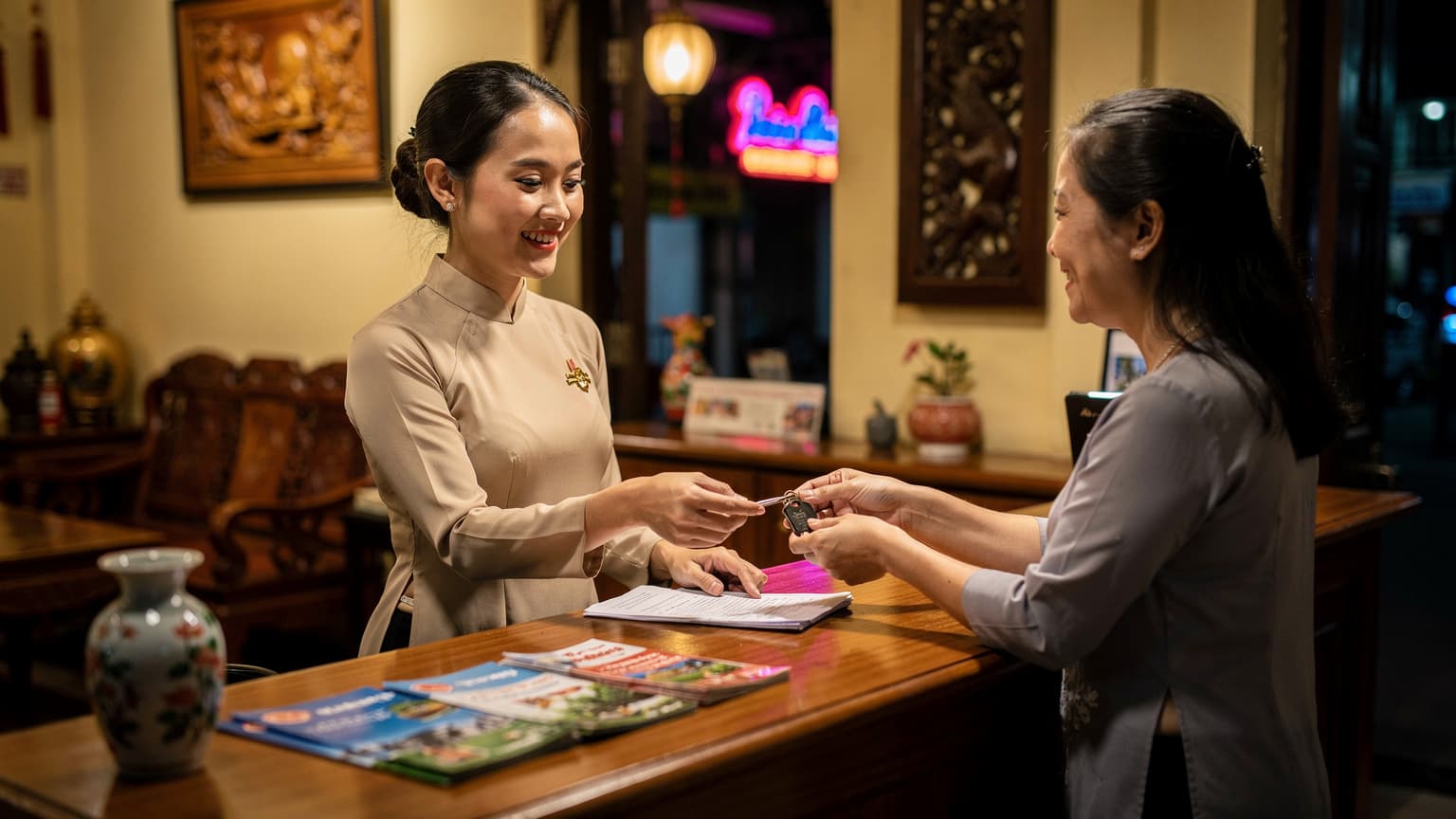 Một nhân viên khách sạn thân thiện đang trao chìa khóa phòng và giấy tờ cho một gia đình Việt Nam tại quầy lễ tân ấm cúng, có thể thấy ánh đèn neon phản chiếu nhẹ nhàng từ bên ngoài cửa sổ nhỏ trên con phố tấp nập của một thành phố Châu Á vào ban đêm.