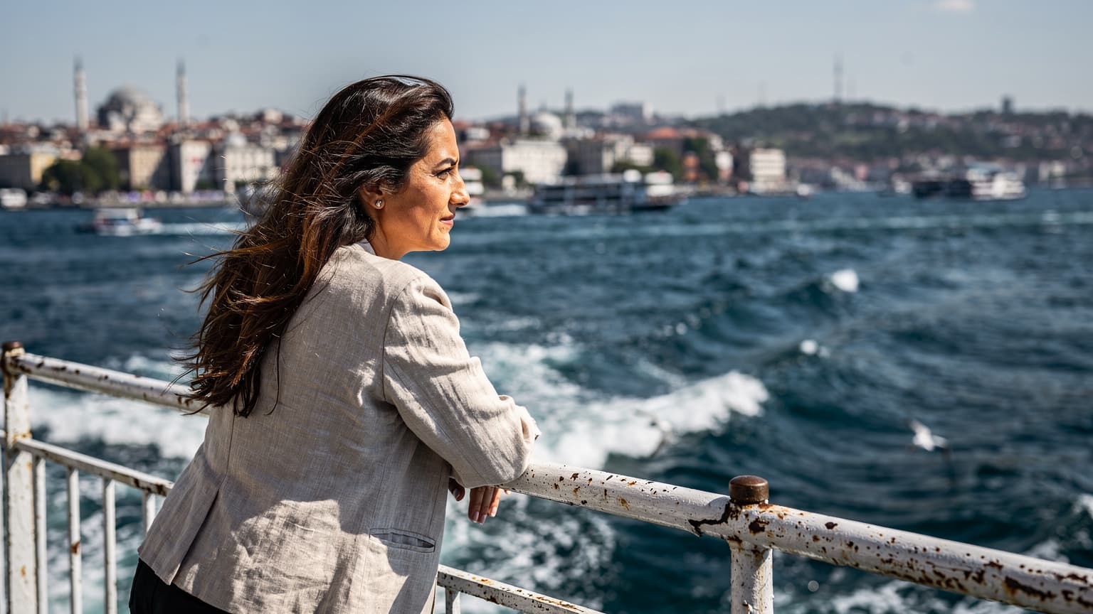 Karaköy'de bir arka sokakta, güneş ışıklarıyla aydınlanmış eski bir binanın önünde, rüzgarda saçları dalgalanan bir kadın vapur iskelesine doğru yürürken, arkasında deniz ve tarihi binaların silüeti görünüyor.