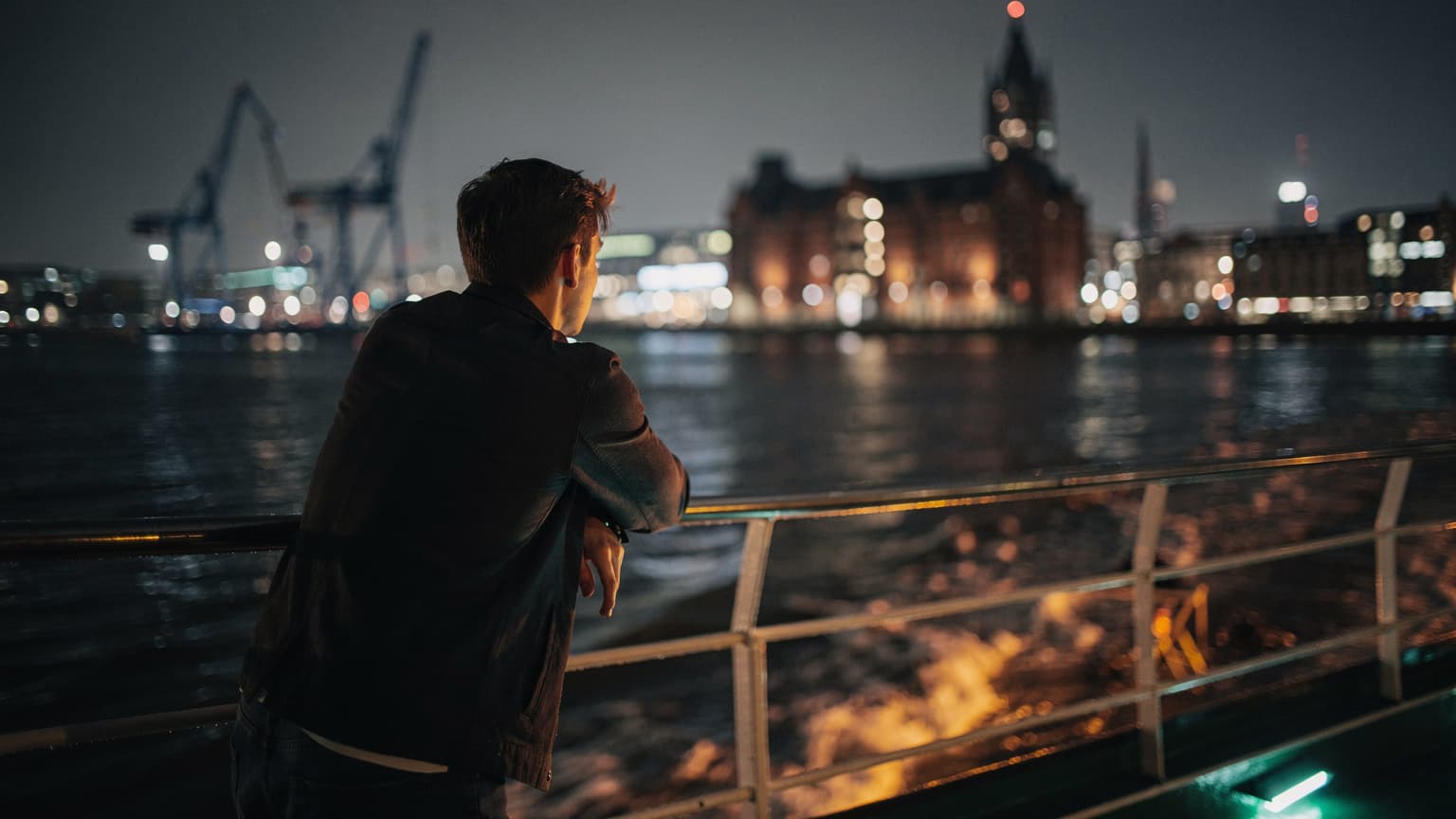 Eine Person lehnt auf dem Deck einer Fähre im Hamburger Hafen bei Nacht, der Wind fährt durch ihr Haar, im Hintergrund sind die beleuchteten Docks und die Skyline zu sehen.