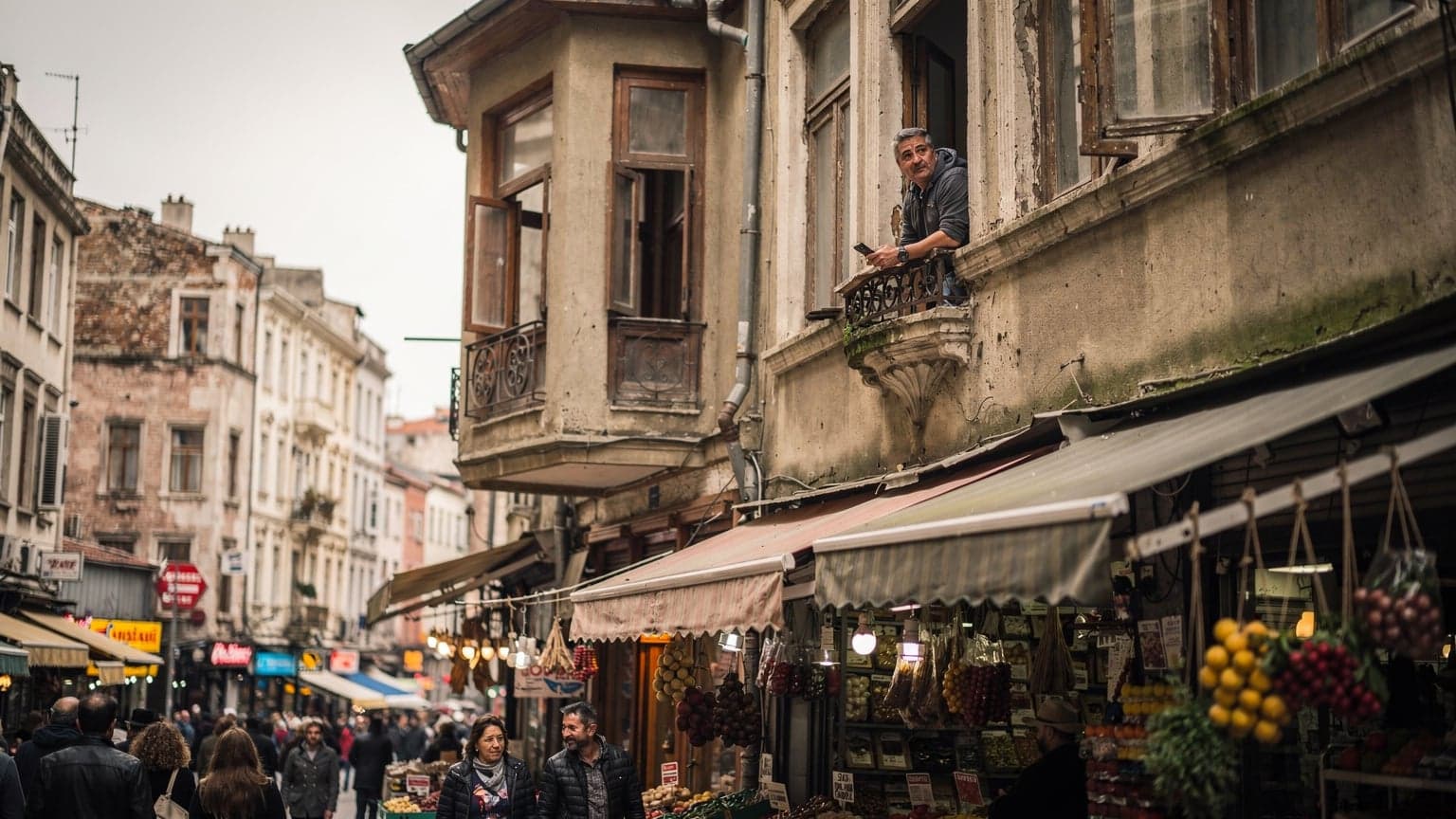 İstanbul Kadıköy çarşısındaki hareketli bir sokağın tepeden görünüşü. Kalabalık caddede yürüyen insanlar ve geleneksel dükkan tabelaları görülüyor. Orta yaşlı bir gezgin, eski bir apartmanın birinci katındaki pencereden sokağı izliyor, elinde bir telefon tutuyor.