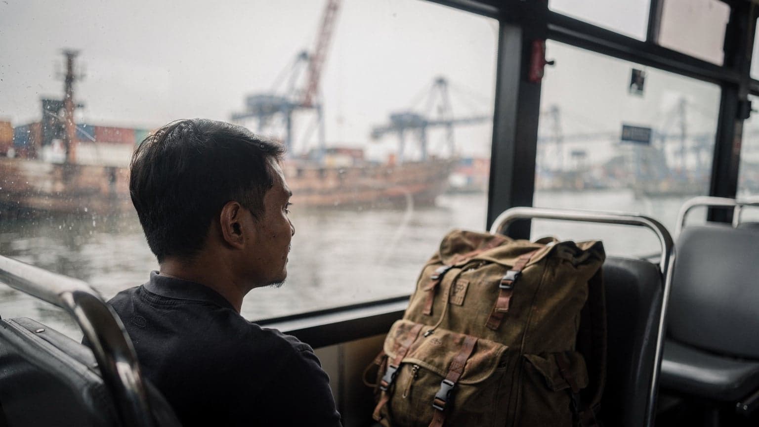 Seorang pelancong duduk di dekat jendela bus di Pelabuhan Sunda Kelapa, Jakarta Utara, tas ransel di kursi sampingnya, cahaya pagi yang lembut masuk dari balik kaca.