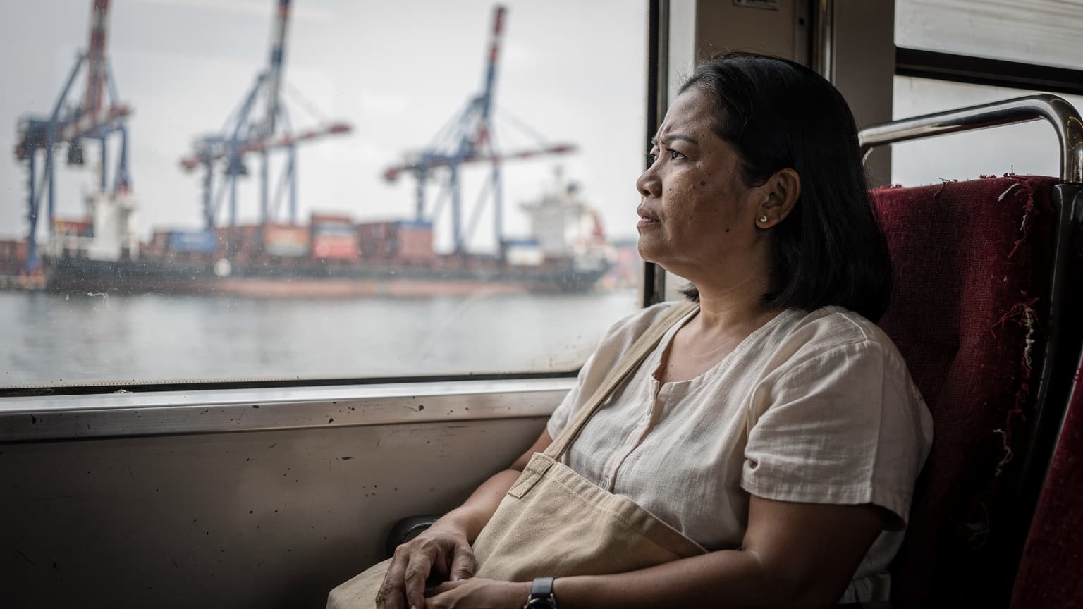 Seorang ibu dengan tas selempang menatap keluar jendela kereta commuter di kawasan Tanjung Priok, Jakarta Utara, langit mendung tipis menyinari pelabuhan di kejauhan