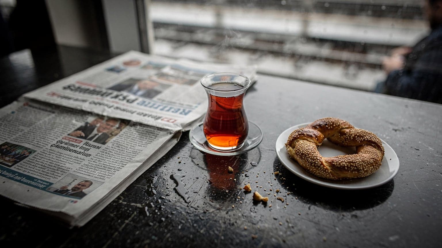 Ankara YHT peronunda yağmurlu gri bir günde, bir masanın üzerinde taze demlenmiş bir fincan çay, yanında açık bir yerel gazete ve ufak bir simit tabağı ile bekleyen bir gezginin üstten çekilmiş fotoğrafı.