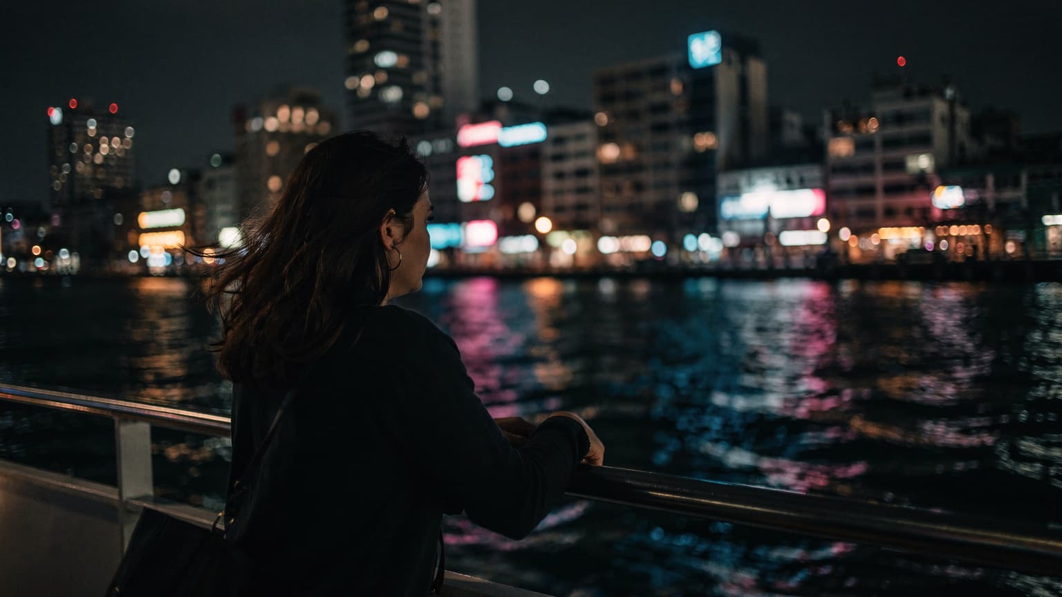 A person with wind in their hair, leaning on the railing of a ferry deck at night, with the illuminated coastline of a foreign city visible in the distance.