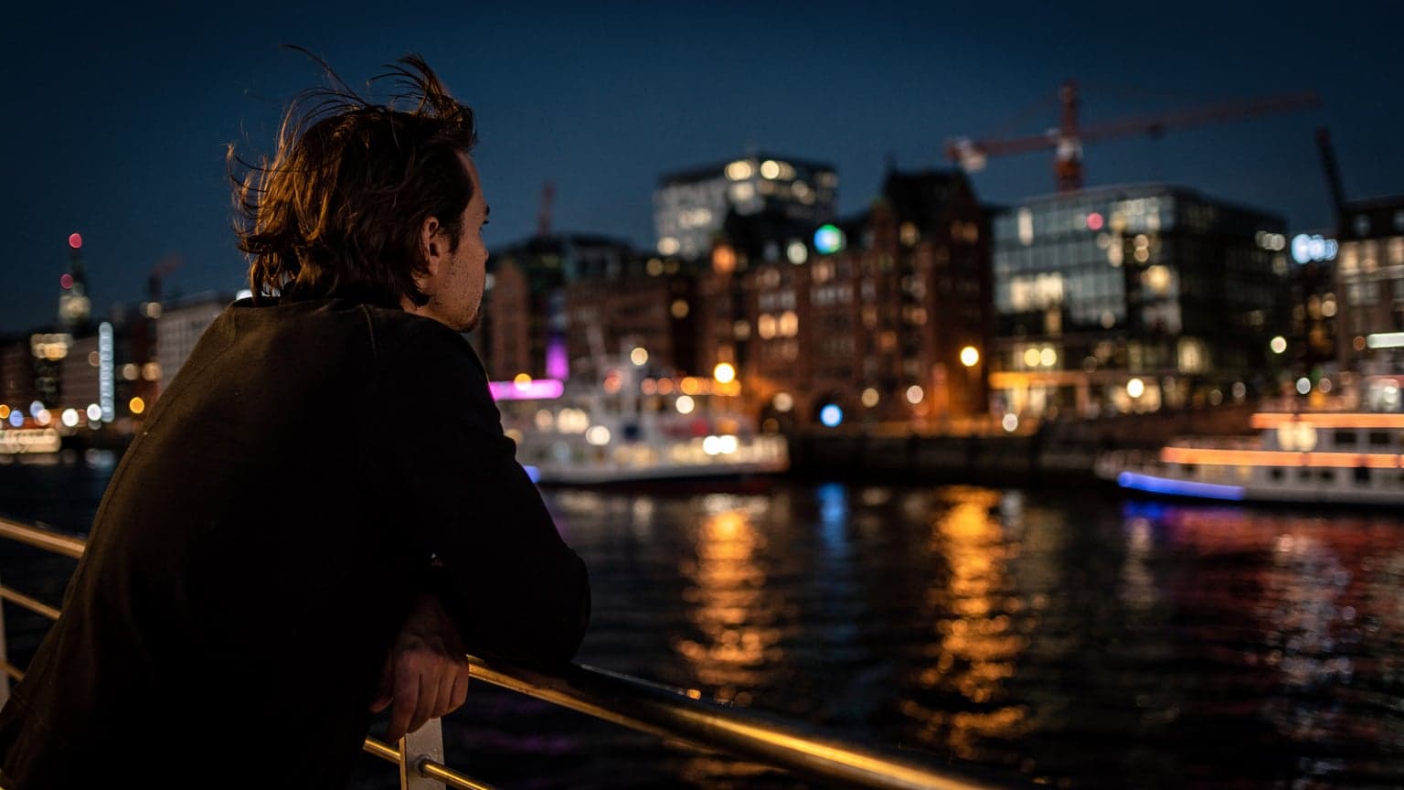 Eine Person lehnt an der Reling einer Fähre bei Nacht, der Wind zerzaust ihre Haare, mit der beleuchteten Hamburger Skyline im Hintergrund.