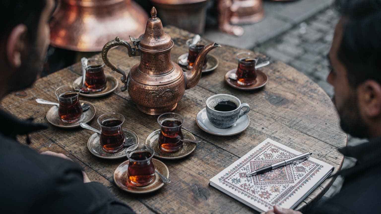 Gaziantep'te bir bakırcı sokağındaki geleneksel bir kafede, masanın üzerinde duran bakır çaydanlık, birkaç bardak demli çay ve yanında taze çekilmiş kahve ile dolu fincanlar. Açık havada, gün ortası ışığı hafif bulutlu bir havadan geliyor, masaya düşen yumuşak gölgeler oluşturuyor. Bir köşede, üzerinde bölgesel motifler olan küçük bir defter ve bir kalem duruyor.