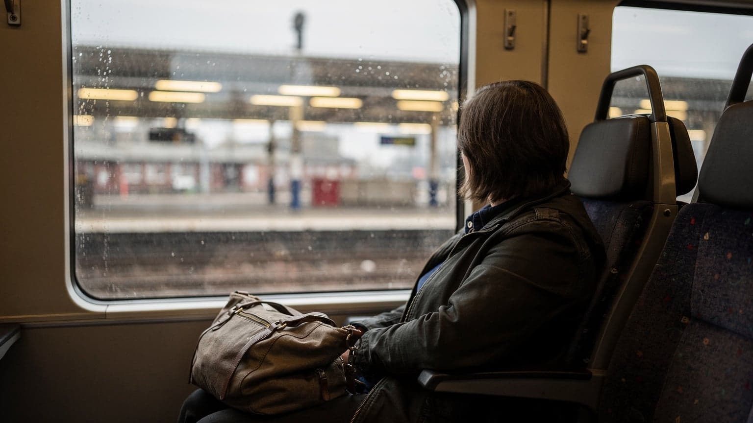 A traveler gazes out a train window at a grey, rainy suburban rail platform, a travel bag resting on the seat beside them, the wet exterior reflecting the interior lights.