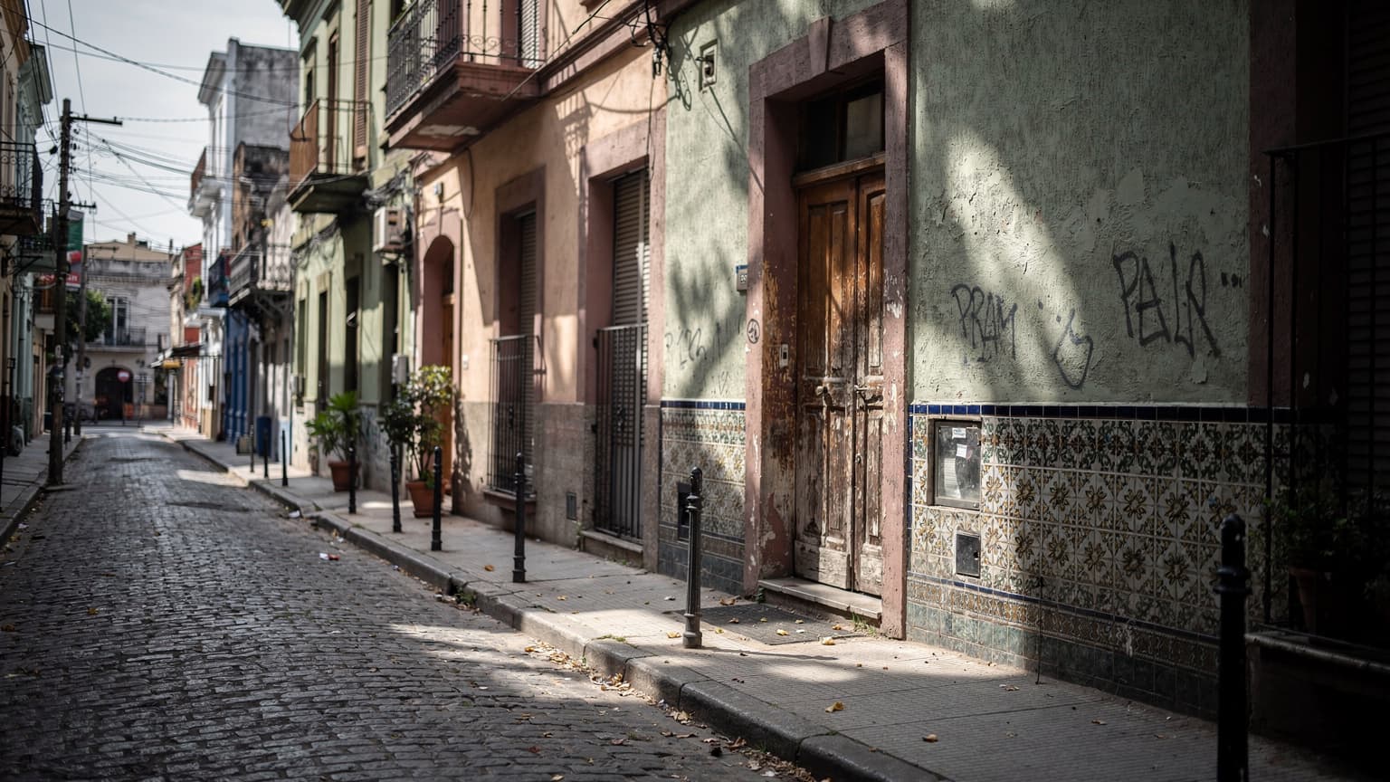A quiet, sun-dappled street in a suburban neighborhood of Buenos Aires, with traditional tile work and a worn wooden door, no people visible.