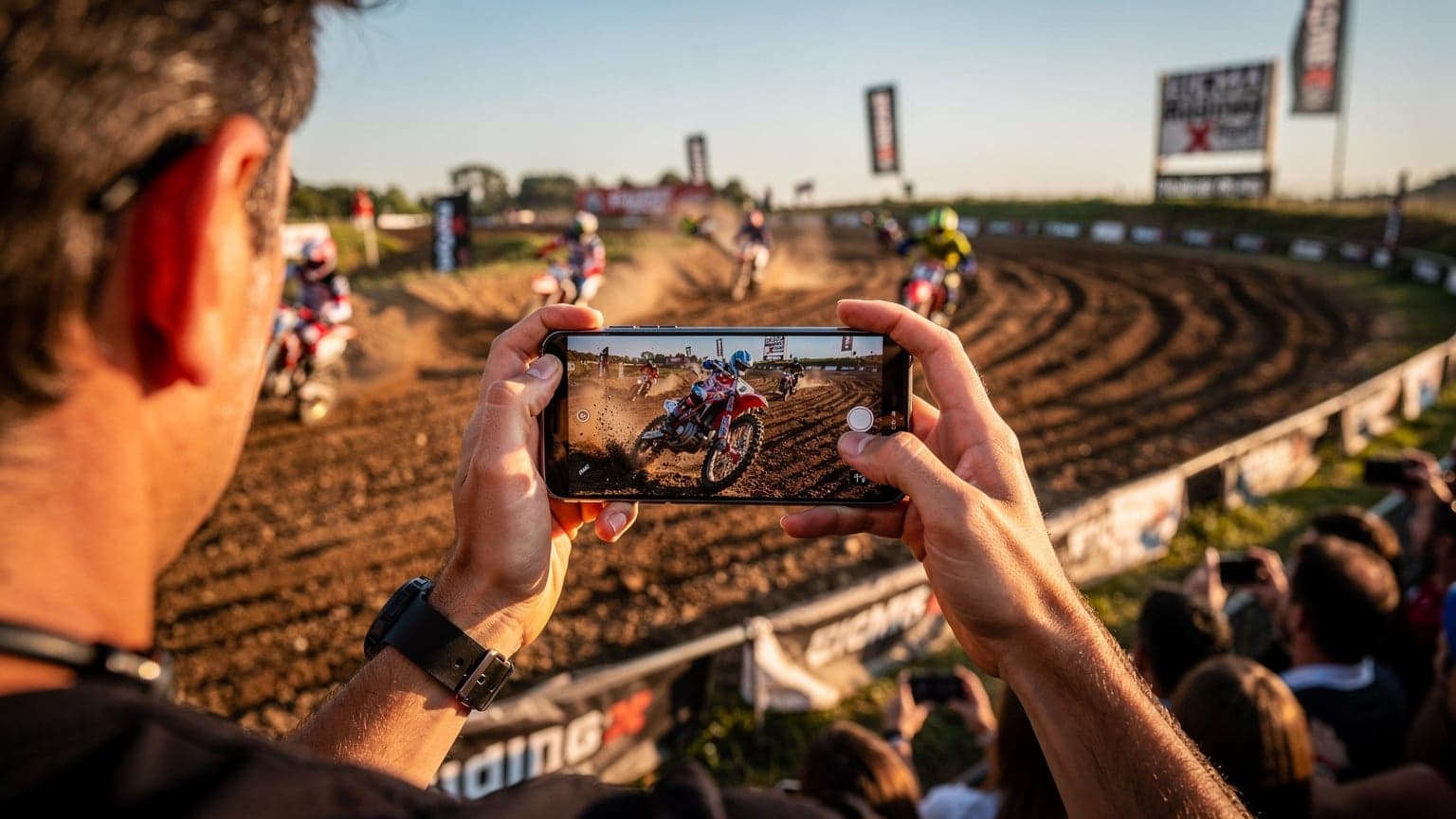 Une personne utilisant son smartphone pour filmer une course de motocross lors de l'EICMA Riding X Fest, avec des motos tout-terrain en arrière-plan et un paysage italien ensoleillé.