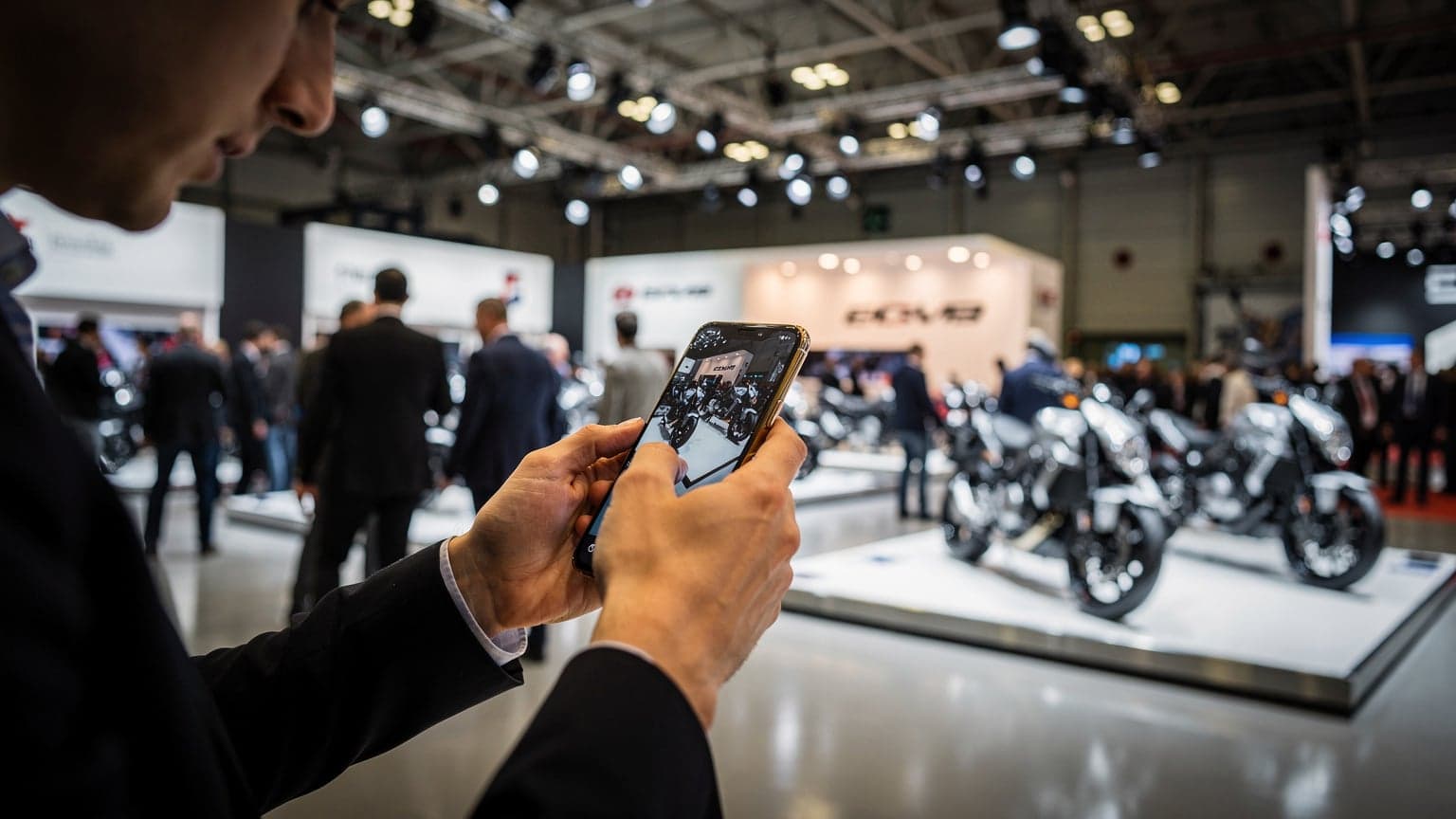 Ένας επισκέπτης της EICMA χρησιμοποιεί το κινητό του τηλέφωνο για να πλοηγηθεί στην έκθεση, με φόντο μοτοσικλέτες και περίπτερα.