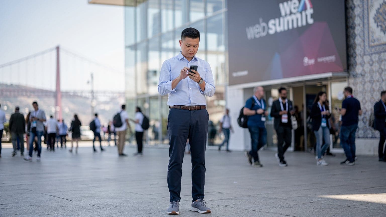 一位中国科技专业人士在里斯本Web Summit会场外使用手机,背景是会议中心和城市风光,象征着高效连接。