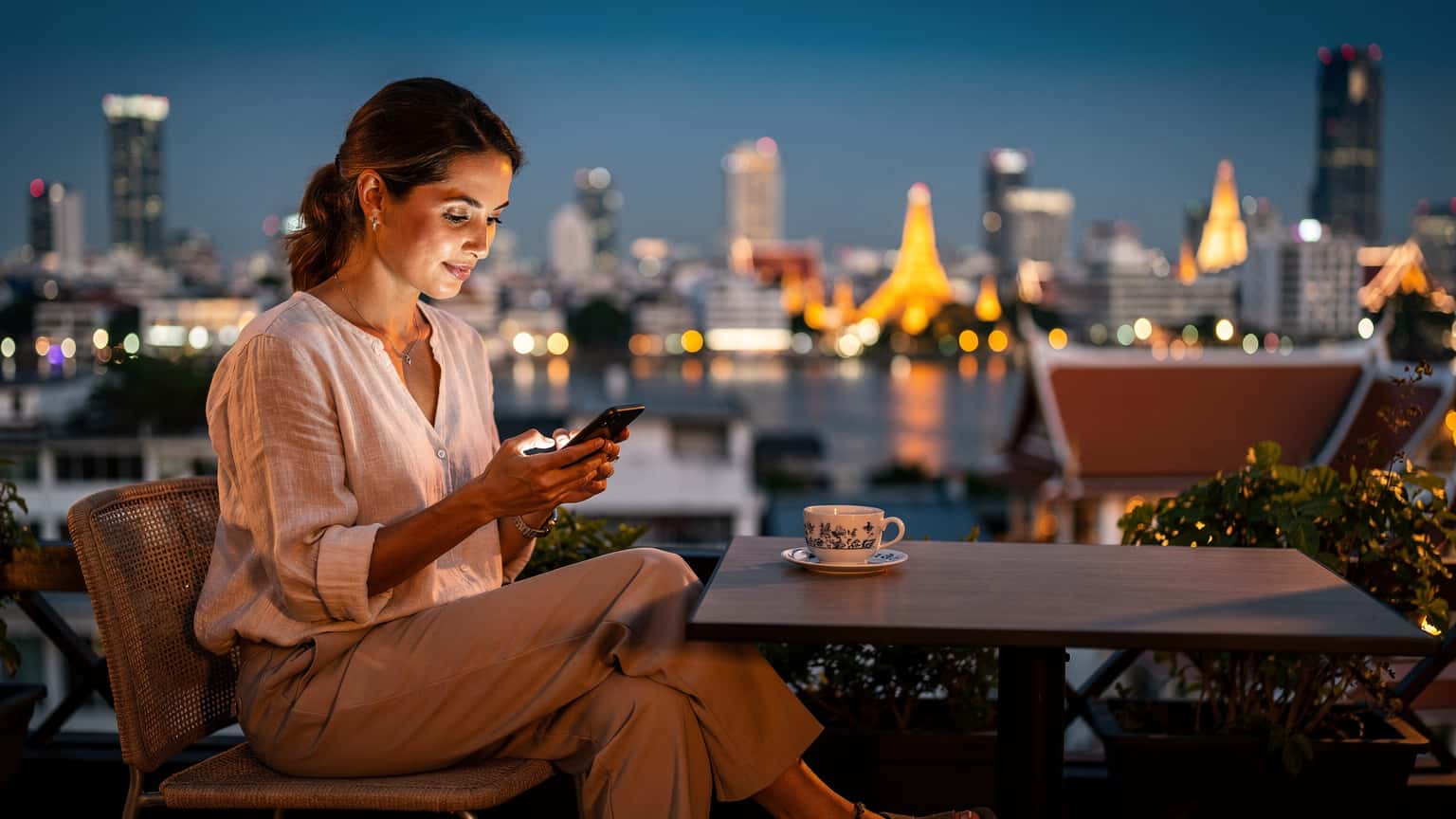 En kvinne i begynnelsen av 30-årene sitter på en takterrasse i Bangkok, ved siden av en tradisjonell thailandsk tekopp, og ser på telefonen sin mens byens skyline med skyskrapere og templer strekker seg ut i det fjerne ved solnedgang.