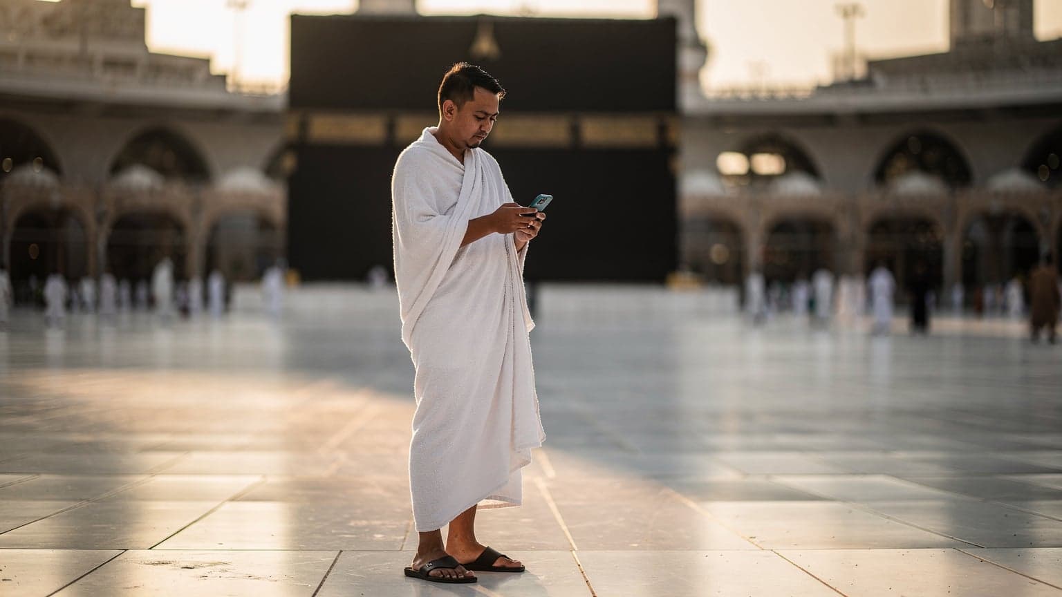 Seorang jamaah haji Indonesia mengenakan ihram, sedang melihat ponselnya di dekat Masjidil Haram di Makkah saat matahari terbit, dengan latar belakang Ka'bah yang indah dan penuh ketenangan.
