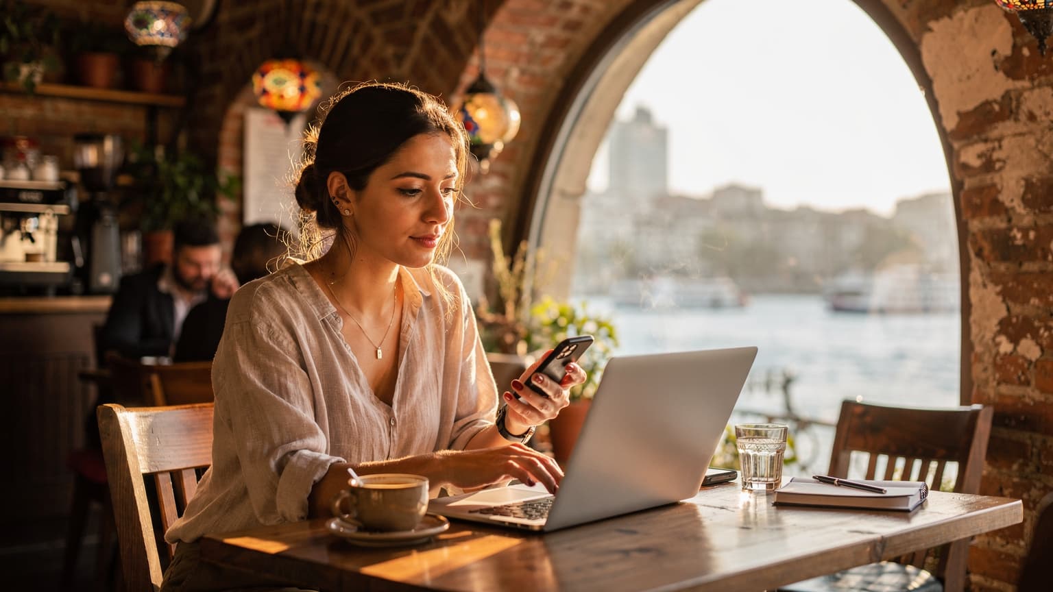 İstanbul'da bir kafede, pencereden gelen altın rengi gün ışığında dizüstü bilgisayarında çalışan ve telefonunda eSIM ayarlarını kontrol eden genç bir kadın.