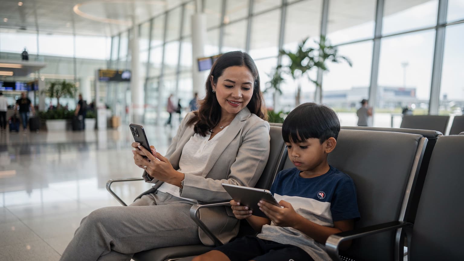 Seorang ibu tersenyum lega sambil memegang Samsung Galaxy S25 di tangan, anaknya di samping sedang asyik menonton tablet di lounge keberangkatan Bandara Changi Singapura. Suasana bandara yang modern dan terang benderang terlihat di latar belakang.