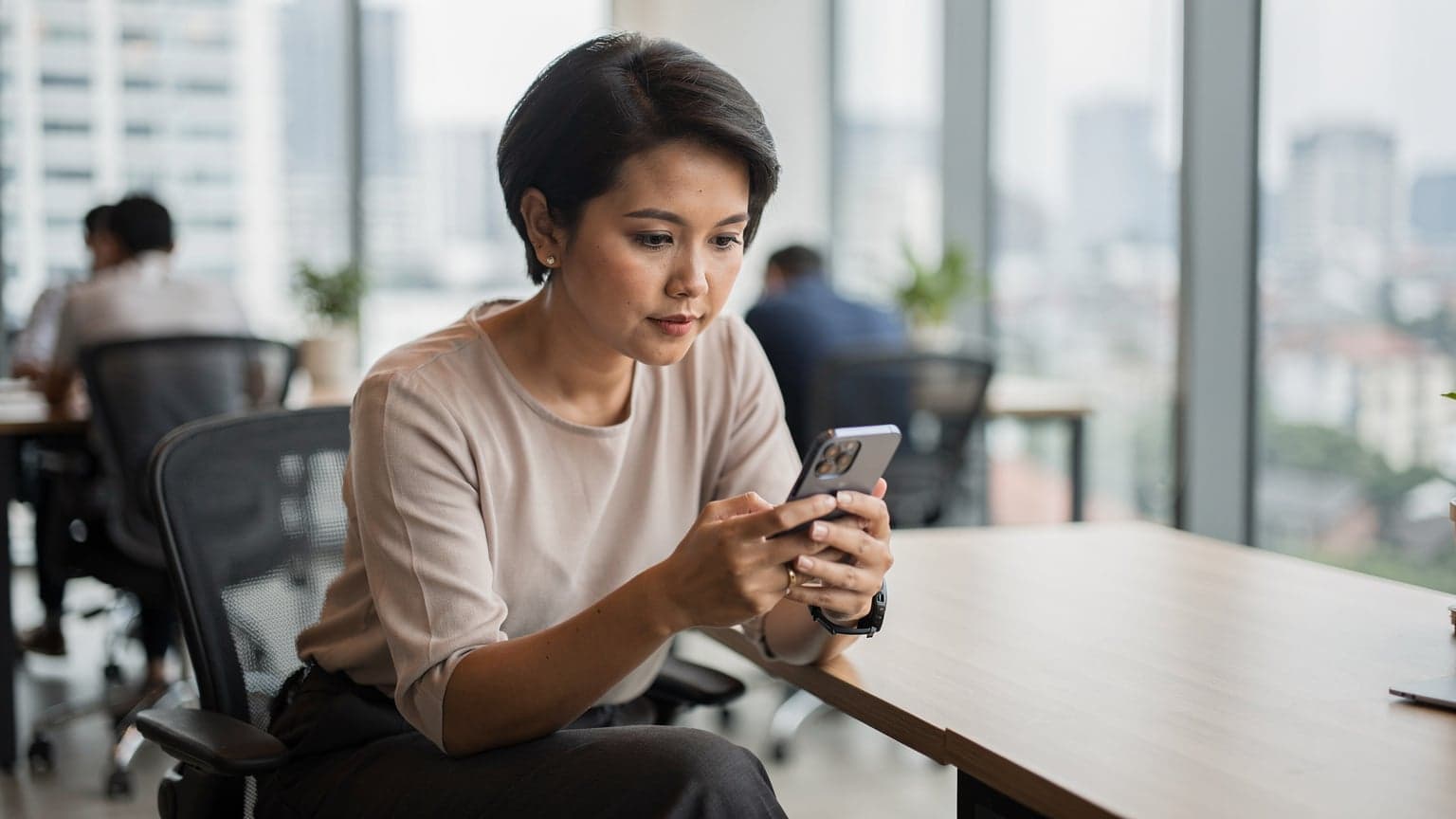 Seorang wanita digital nomad sedang memeriksa ponselnya di sebuah co-working space yang modern di Jakarta, dengan latar belakang jendela besar yang menampilkan pemandangan kota.