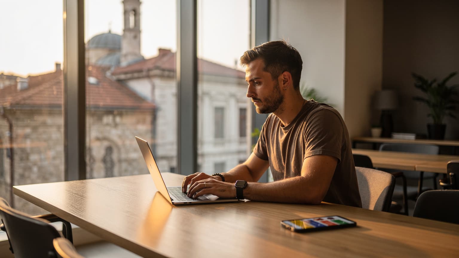 Een jonge digitale nomade zit geconcentreerd in een modern co-working café in Skopje, Noord-Macedonië, met een laptop en een smartphone naast een kopje koffie, genietend van de ochtendzon die door het raam valt.