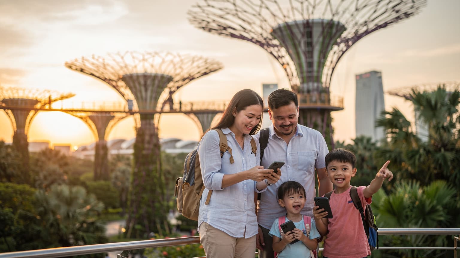 Một gia đình gồm bố mẹ và hai con nhỏ đang đi bộ qua Supertree Grove tại Gardens by the Bay, Singapore vào buổi tối, với những cây siêu cây phát sáng rực rỡ phía sau.