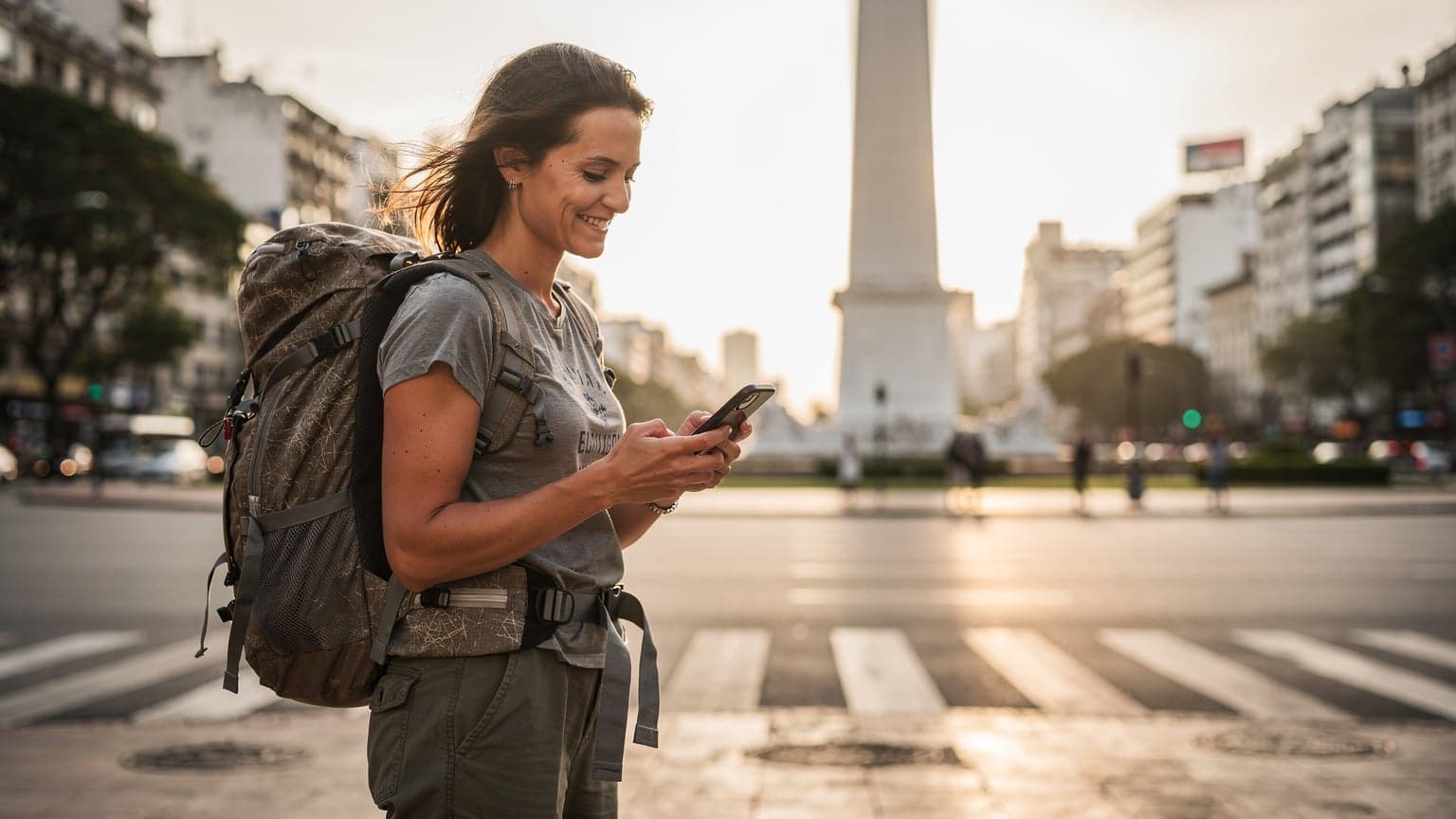 Uma mochileira sorridente, com uma mochila grande, segurando um smartphone com um mapa aberto, em frente ao Obelisco de Buenos Aires ao pôr do sol.