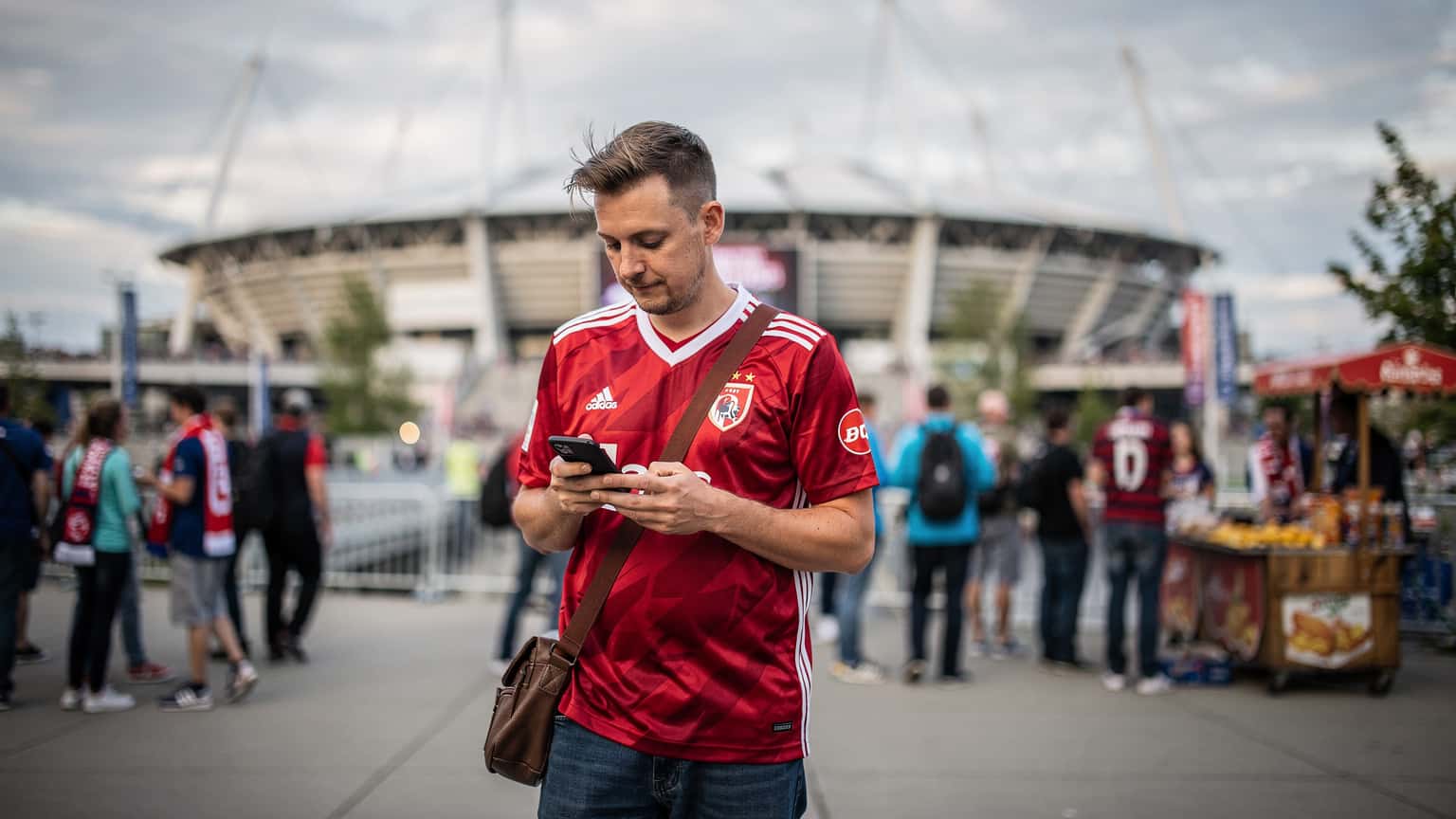 En fotballfan i en landslagsdrakt, med en mobiltelefon i hånden, navigerer et travelt stadionområde i Vancouver, Canada, med BC Place i bakgrunnen.