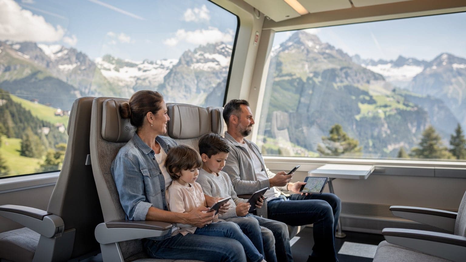 Eine Familie, bestehend aus Eltern und zwei Kindern im Alter von 8 und 11 Jahren, sitzt in einem Panoramazug, der durch die Schweizer Alpen fährt. Die Kinder schauen auf ihre Tablets, während die Eltern die vorbeiziehende Landschaft bewundern und ein Smartphone in der Hand halten, das eine Karte anzeigt.