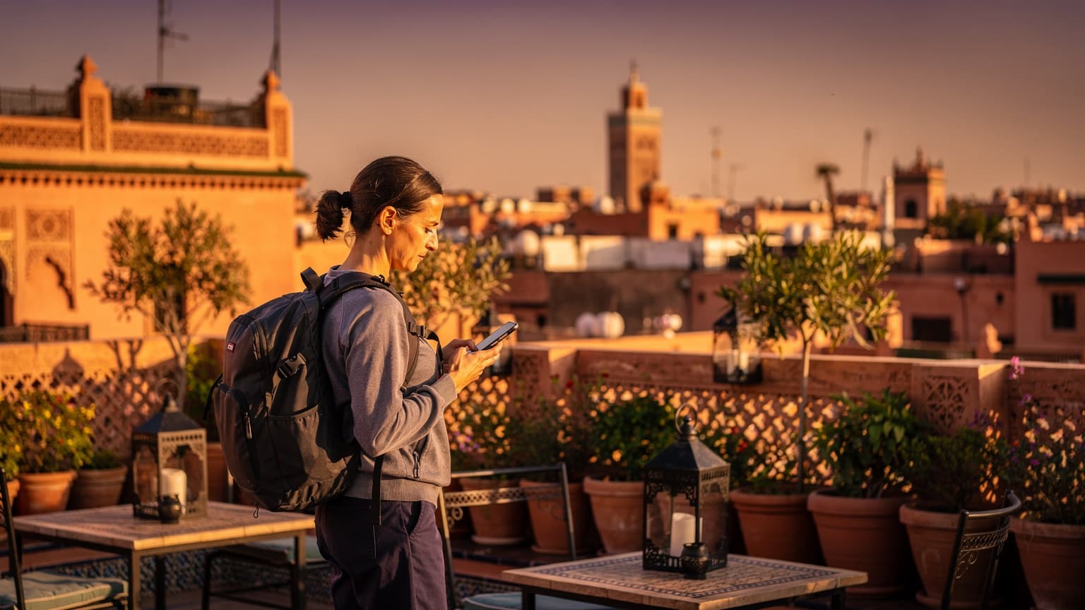 Een reiziger kijkt op een smartphone bij zonsondergang over de daken van Marrakech, met de Koutoubia-moskee op de achtergrond.