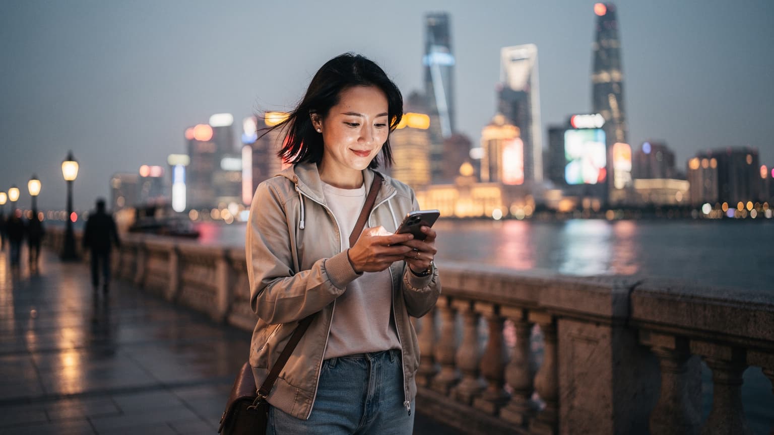 中国の上海にある外灘の夜景を背景に、スマートフォンを操作してeSIMの設定を確認している日本人旅行者。